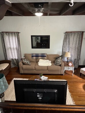 Uma sala de estar com sofá bege, TV fixada na parede, ventilador de teto e mesa de centro de madeira.