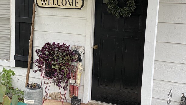 Una puerta negra con un cartel que dice 'WELCOME' encima, una corona colgada en la puerta y un pequeño jardín con plantas y un barril metálico.