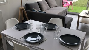 Uma área de refeições com uma mesa branca posta para quatro pessoas, um sofá cinzento e vista para um jardim através de uma porta de vidro.