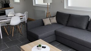 Uma sala de estar moderna com um sofá cinzento, uma mesa de centro de madeira e uma área de jantar com uma mesa e cadeiras brancas.