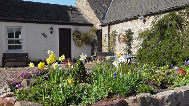 A stone building with a black door, a bench, and a garden with various flowers. - Perfectly located for The Pentlands. It also has easy access to the city. (Edinburgh)