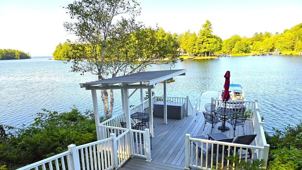 En træterrasse med pergola, udendørs møbler og udsigt til en sø.
