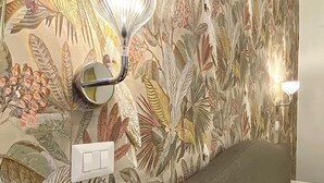 Een kamer met bloemen behang, een bed met een wit kussen, en een wandlamp.