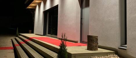 A modern building with a red carpet leading to a wooden bench area, a small tree, and a gravel path.
