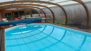 En swimmingpool med træterrasse og glasvægge, omgivet af et drivhuslignende bygning.