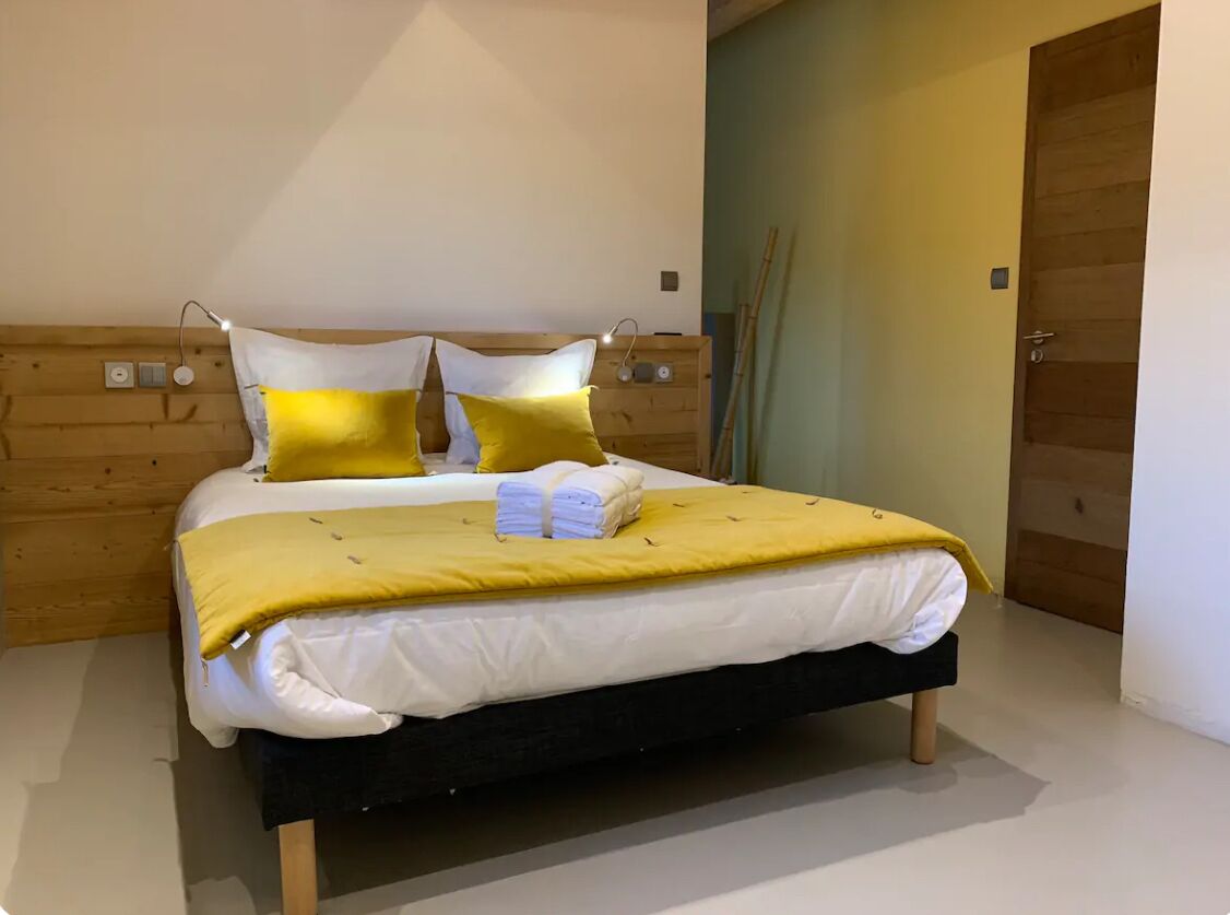 Una cama bien hecha con una manta y almohadas amarillas, un cabecero de madera y una mesita de noche con una lámpara.