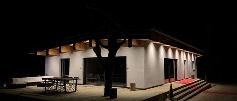 A modern building with a flat roof, a covered outdoor area with a table and chairs, and a red carpet leading to the entrance.