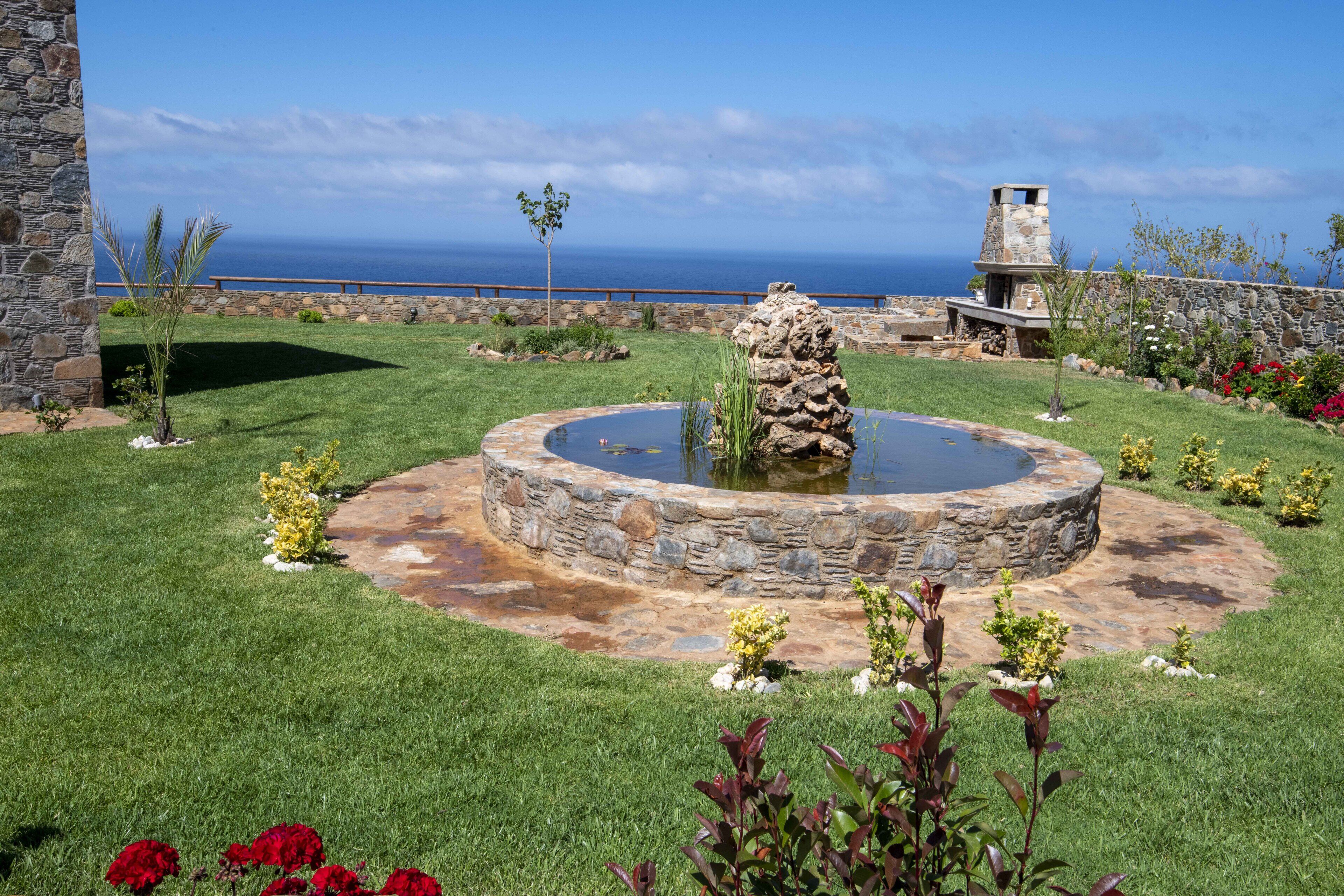 Un jardín ajardinado con un estanque circular de piedra, un muro de piedra y vistas al océano.