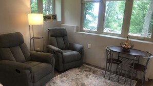 Een woonkamer met twee grijze fauteuils, een vloerlamp, een klein rond tafeltje met een draadmand, en uitzicht op bomen door de ramen.