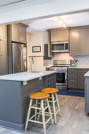 A modern kitchen with grey cabinets, stainless steel appliances, a marble countertop, and two wooden stools.
