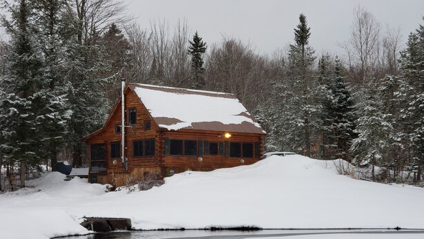 Une cabane en rondins dont le toit est recouvert de neige, entourée d’arbres et bordée par une étendue d’eau gelée.