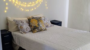 Une chambre à coucher avec un lit, deux oreillers, une table de chevet, une lampe fixée au mur et un plancher en bois.
