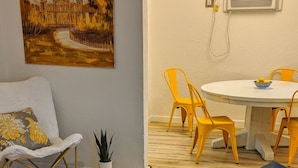 Un salon avec un canapé bleu foncé, des coussins jaunes et blancs, un tapis à motifs rouges, un tableau représentant une église, un coin repas avec une table blanche et des chaises jaunes, et une plante en pot.