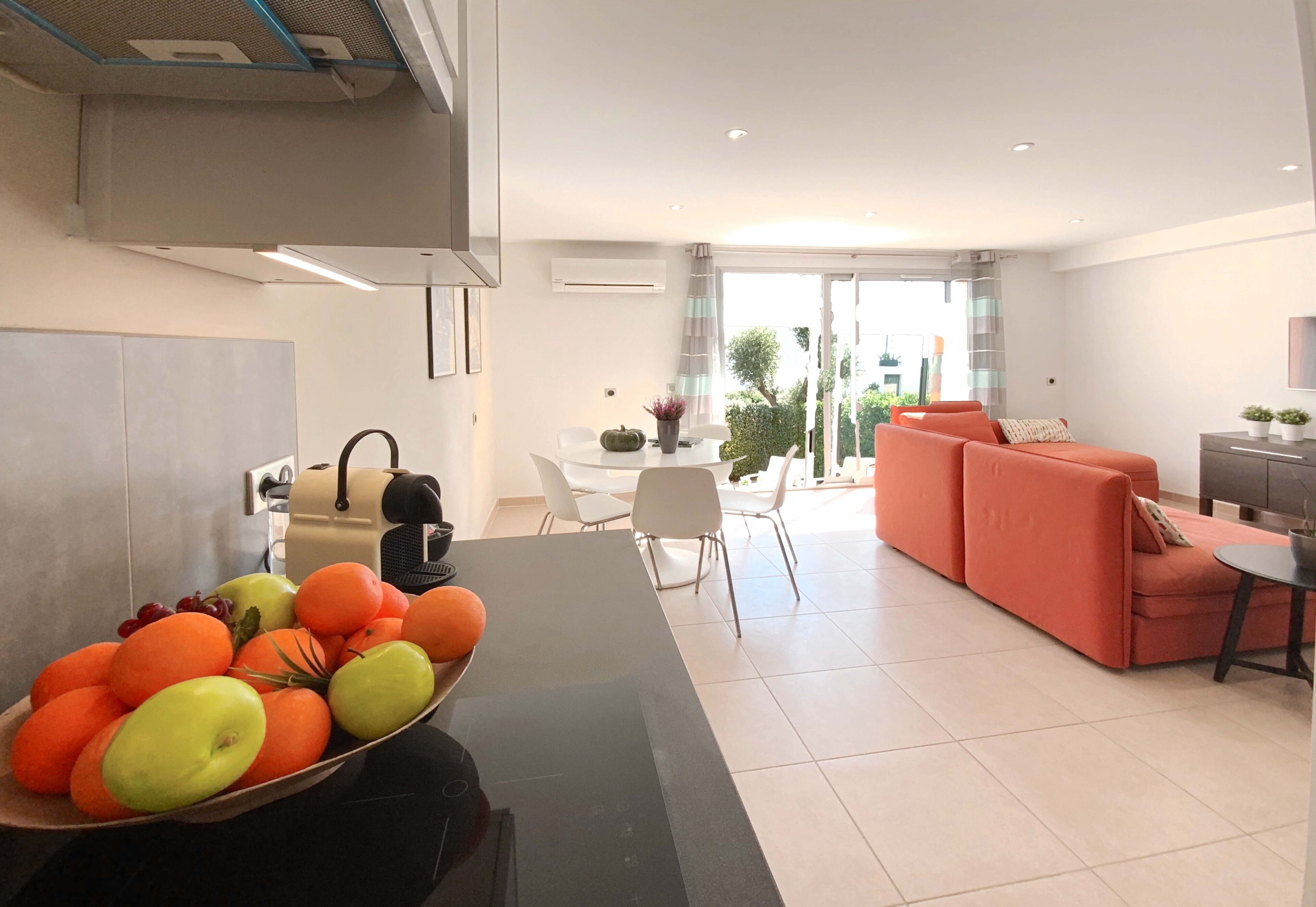 Une cuisine moderne avec un panier de fruits, un coin repas avec une table et des chaises blanches, et un salon avec un canapé et un téléviseur.