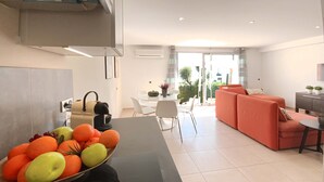 Een moderne keuken met een fruitschaal, een eethoek met een witte tafel en stoelen, en een zithoek met een bank en een televisie.