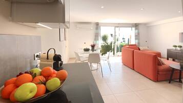 Een moderne keuken met een fruitschaal, een eethoek met een witte tafel en stoelen, en een zithoek met een bank en een televisie.