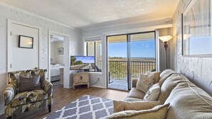 Una sala de estar con sofá, sillón y televisor, un balcón con vistas, y una puerta que da a un dormitorio.