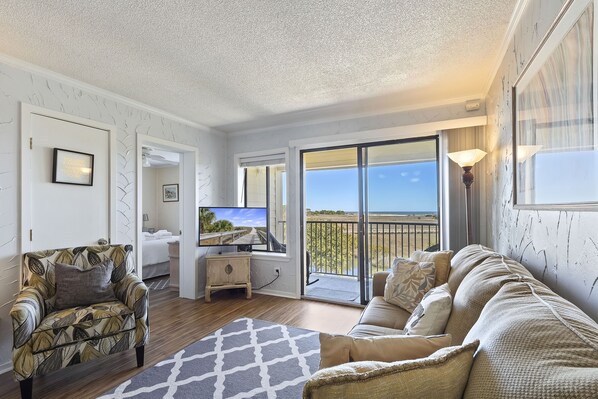 A living room with a sofa, armchair, and TV, a balcony with a view, and a door leading to a bedroom.