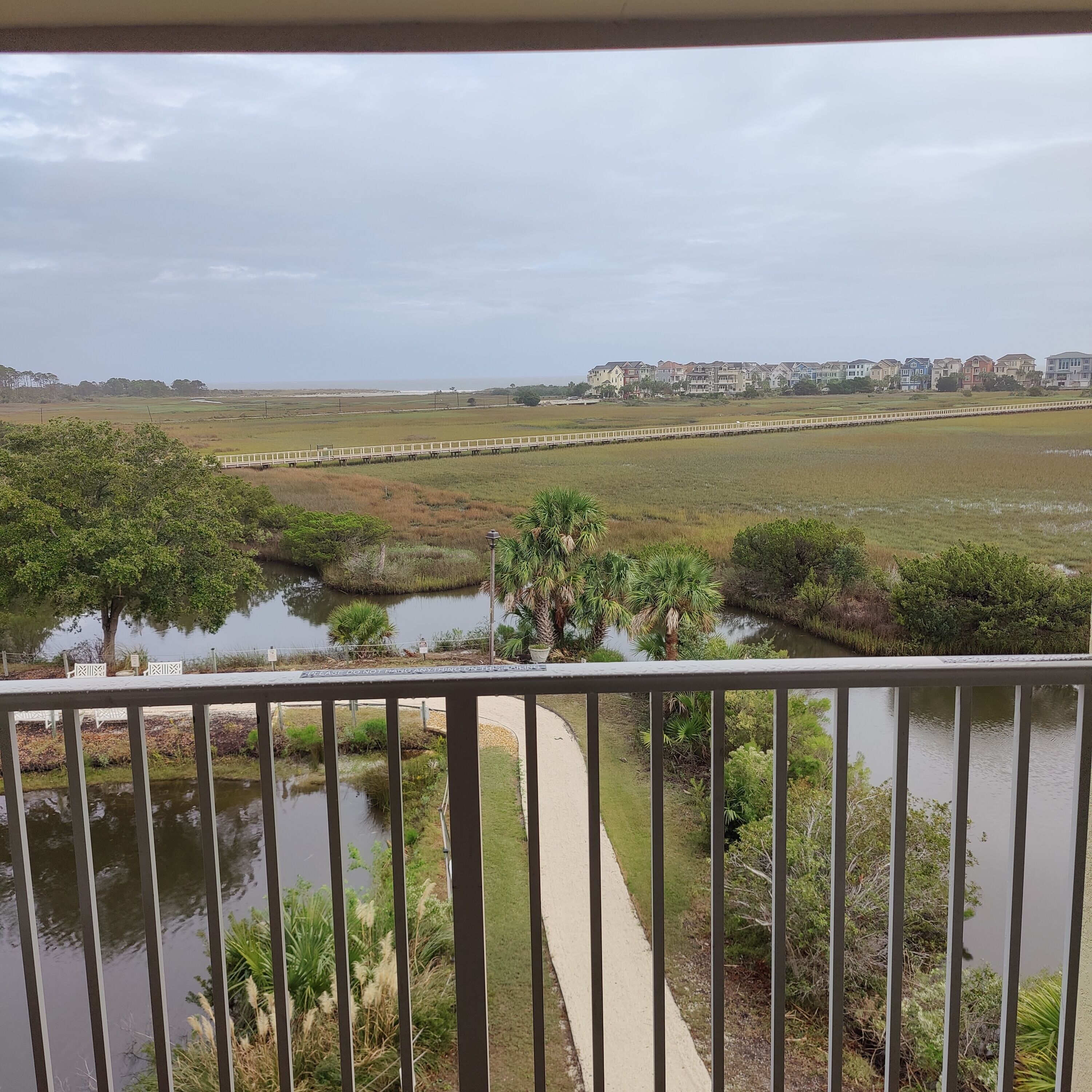 Vue depuis un balcon donnant sur une zone marécageuse, avec un sentier, de petits étangs et, au loin, une rangée de maisons aux couleurs vives.