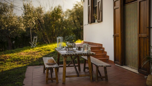 An outdoor patio with a wooden table and benches, surrounded by greenery and a mountain backdrop.