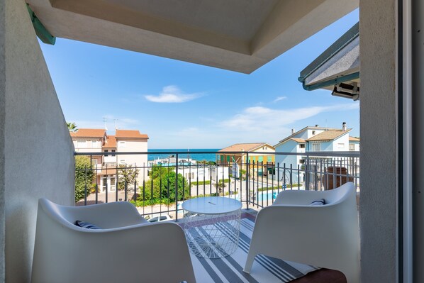 A balcony with two white chairs, a small round table, and a view of the sea and buildings.