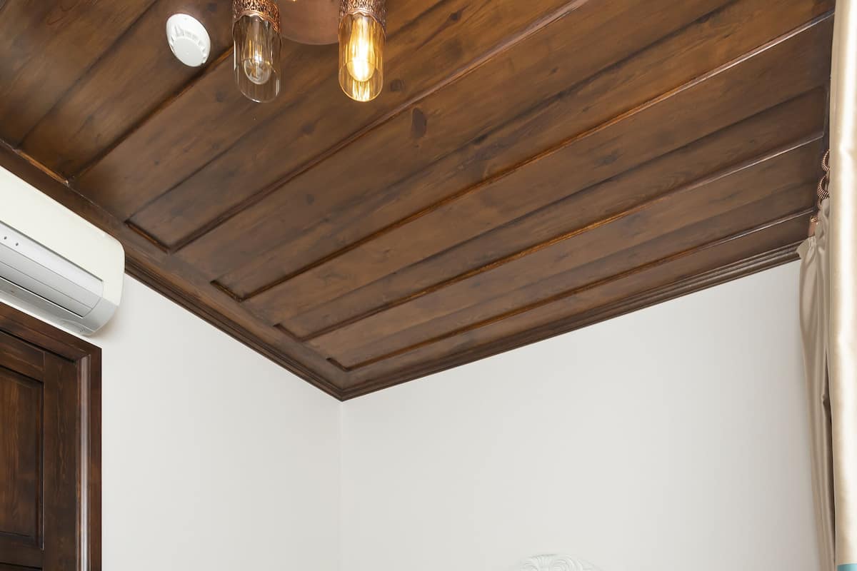 A bedroom with a wooden ceiling, a bed with teal and beige bedding, a white headboard, a nightstand, and a white air conditioning unit.