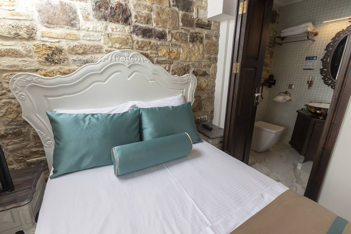 A bed with a white headboard and teal pillows in a room with a stone wall and a bathroom visible through an open door.