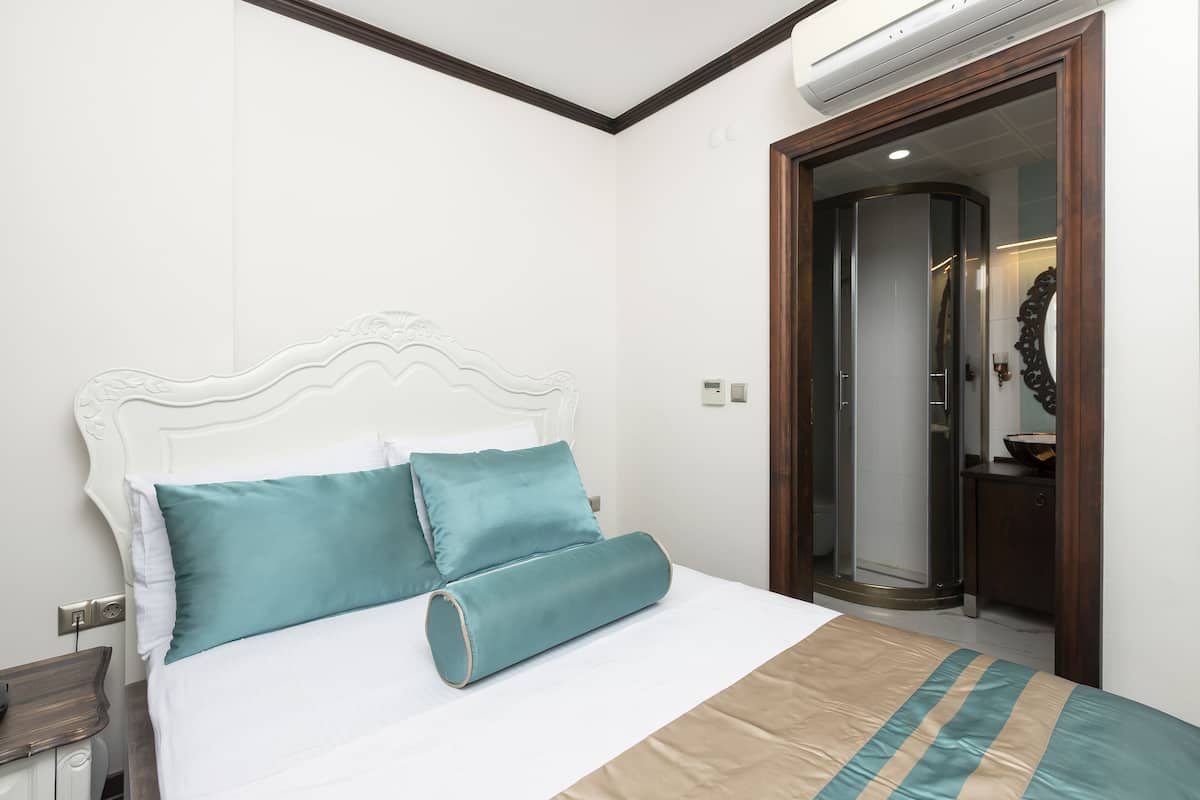 A neatly made bed with a white headboard and teal pillows, a wooden door to the right, and a small bedside table with a lamp.