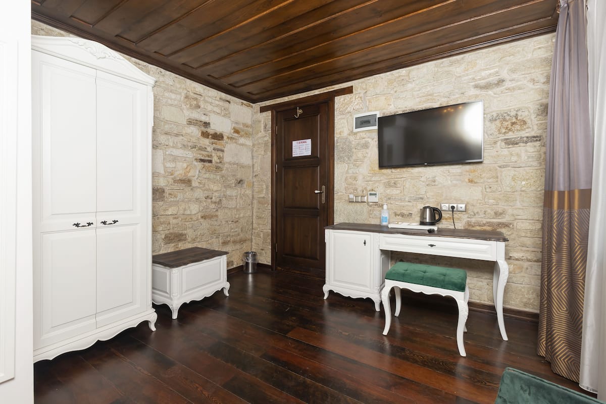 A room with a stone wall, wooden flooring, a white wardrobe, a desk with a green bench, and a flat-screen TV mounted on the wall.