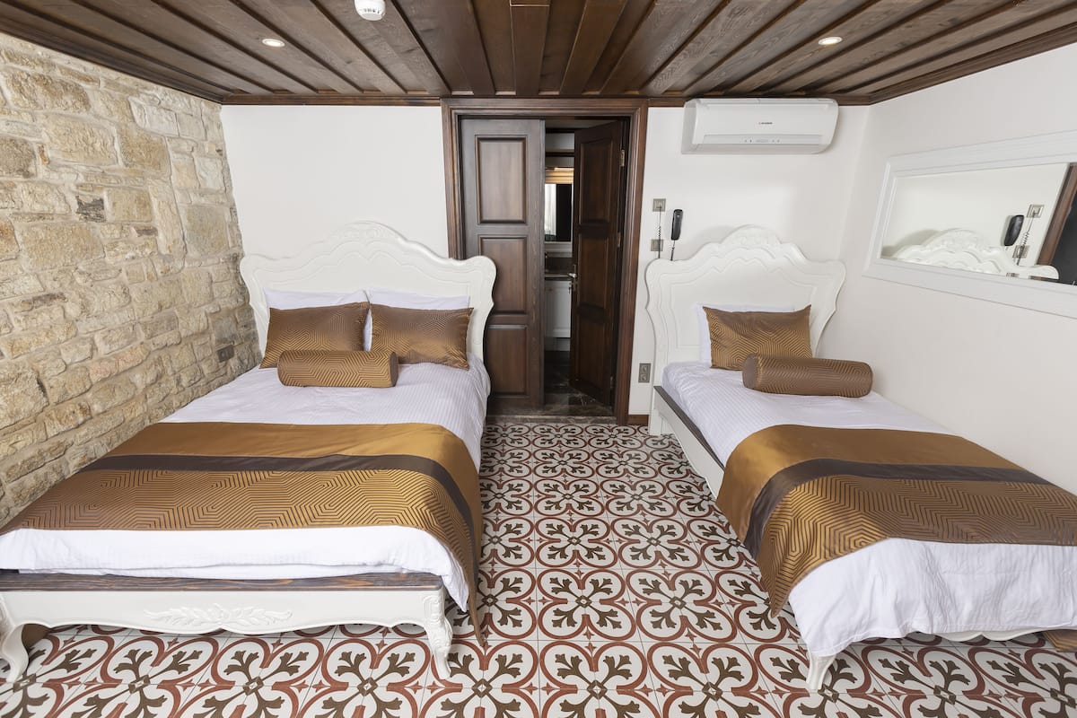 A hotel room with two beds, patterned floor tiles, a stone wall, wooden ceiling beams, and a small kitchenette.