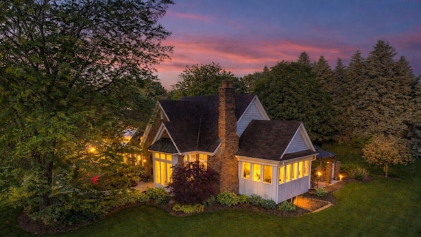 A two-story house with a stone chimney, a small attached structure, and a well-lit garden.