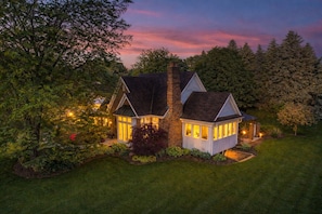 A two-story house with a stone chimney, a small attached structure, and a well-lit garden.