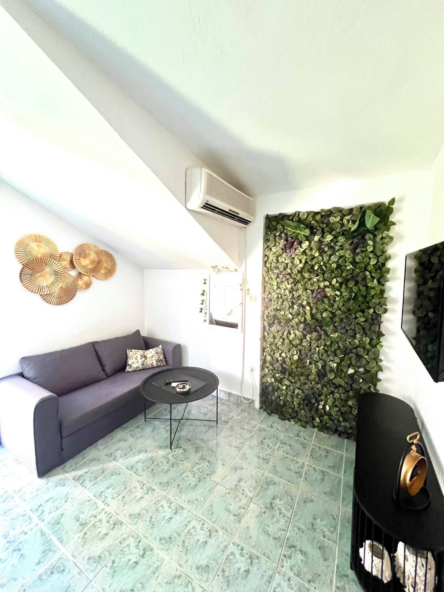 A living room with a grey sofa, a small round table, a wall-mounted air conditioner, and a green leafy wall decoration.