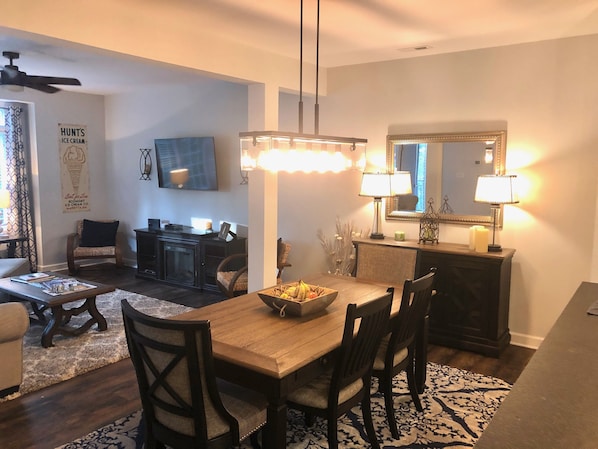 A dining area with a wooden table, four chairs, a chandelier, and a mirror above the table.