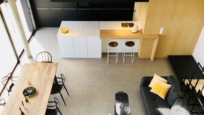 Una cocina moderna con armarios blancos, un área de comedor con una mesa y sillas de madera, y un sofá negro con cojines estampados.