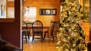 A cozy dining area with wooden walls, a wooden table, and four chairs. A decorated Christmas tree is visible in the background. - "Back-Porch Sittin'" Perfect! Walk-in Cedar Cabin near 76 Country Blvd Strip! (Branson)