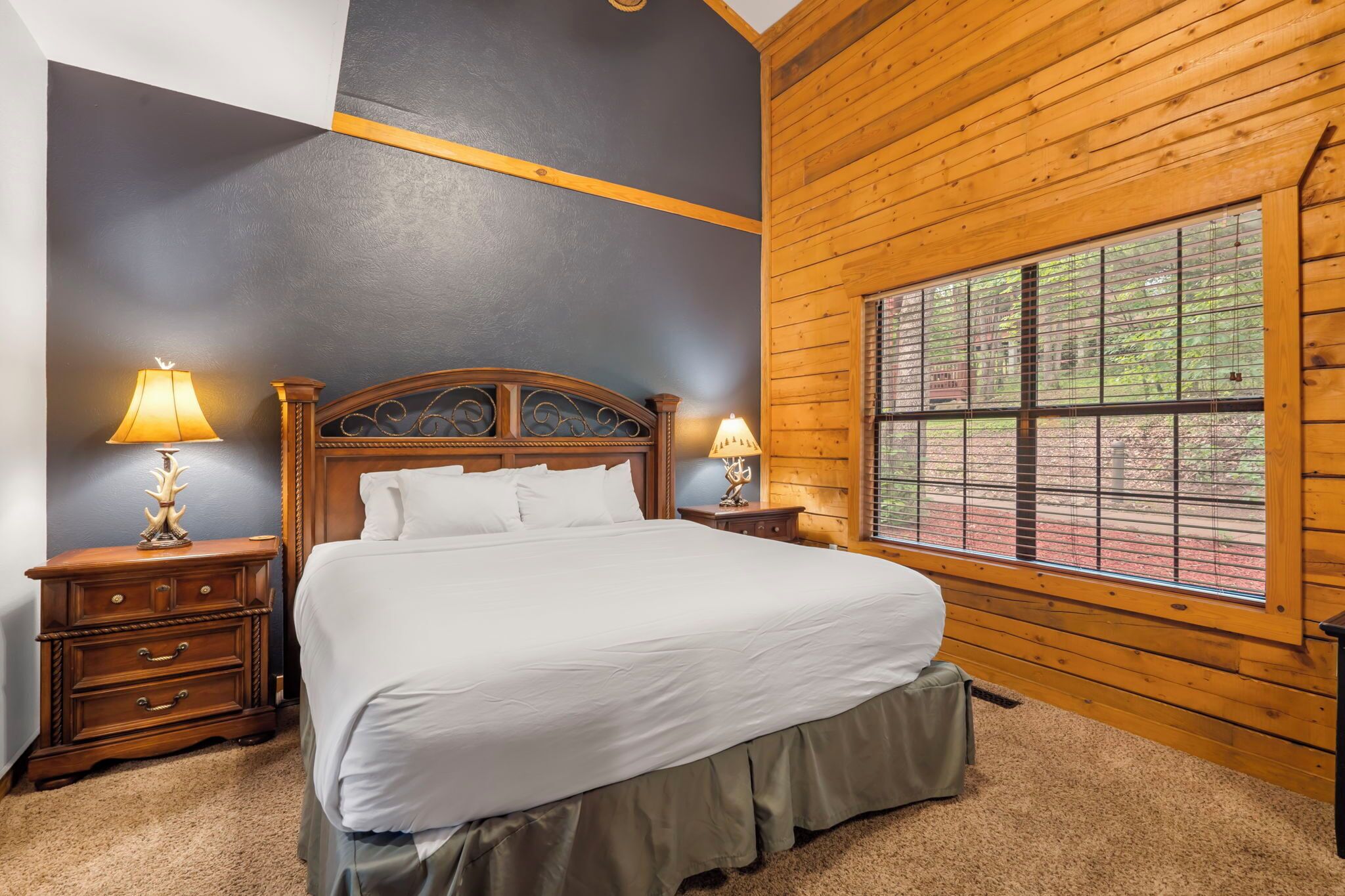 A bedroom with a large bed, wooden headboard, two bedside tables with lamps, a window with a view of the outdoors, and a carpeted floor.
