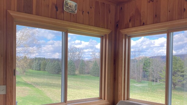 Ein Zimmer mit Holzwänden und -decke, einem großen Ecksofa und zwei großen Fenstern mit Blick auf ein Wiesfeld und Bäume.