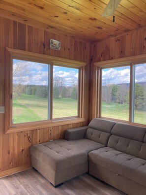 A room with wooden paneled walls and ceiling, a large sectional sofa, and two large windows offering a view of a grassy field and trees. - 1500 sqft private entrance with beautiful views of the Berkshire Mountains (Lee)