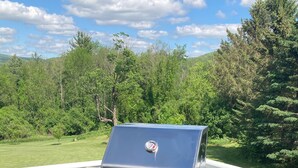 Ein tragbarer Gasgrill auf einer Terrasse mit weißer Geländer, umgeben von Grünpflanzen und Hügeln.