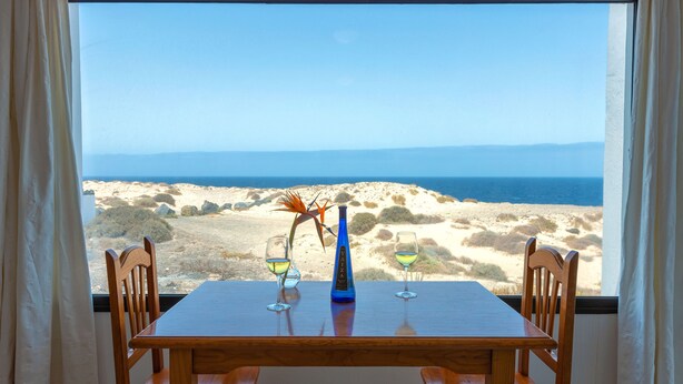 A dining table with two chairs, a bottle of wine, and two wine glasses. The table is set against a view of the ocean and sandy landscape. - Reyes Natur - Bungalow on the seafront (Charco del Palo)