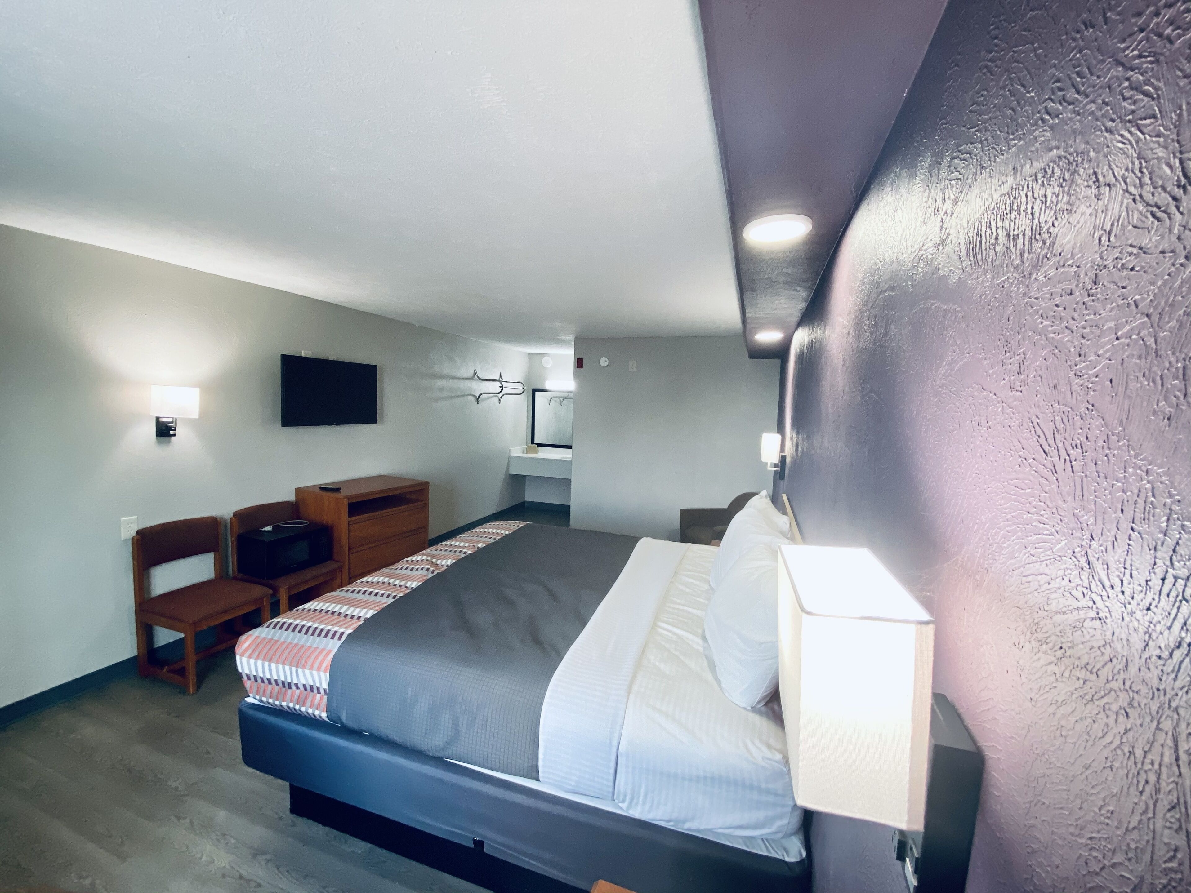 A modern hotel room with a large bed, a desk, a chair, a flat-screen TV, and a bathroom with a mirror and sink.