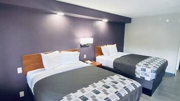 A hotel room with two beds, each with a wooden headboard and white bedding. There is a nightstand with a phone on each bed. The walls are painted purple, and there is a light fixture above the beds.