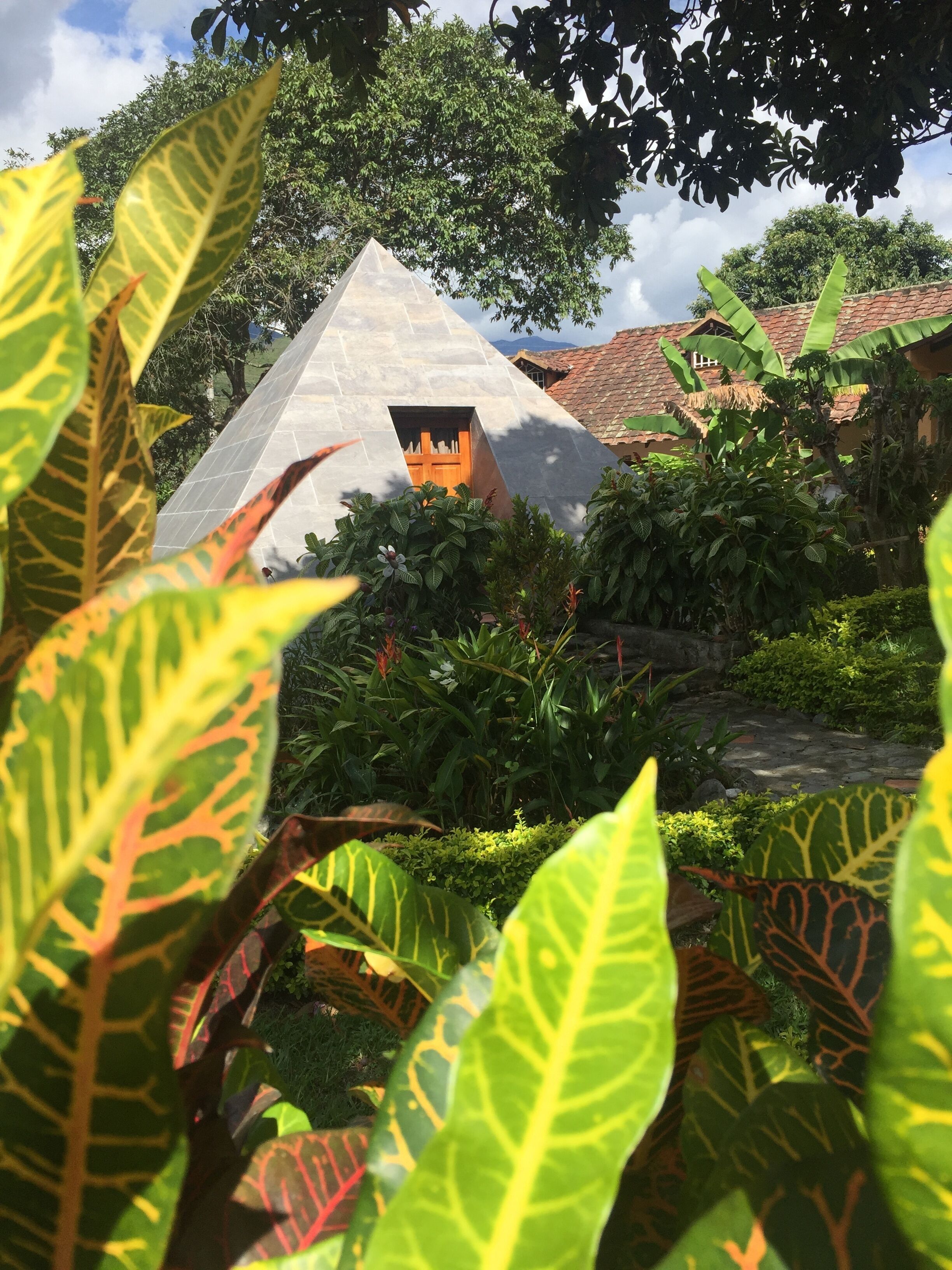 A pyramid-shaped structure with a wooden door, surrounded by lush greenery and a thatched-roof building in the background.
