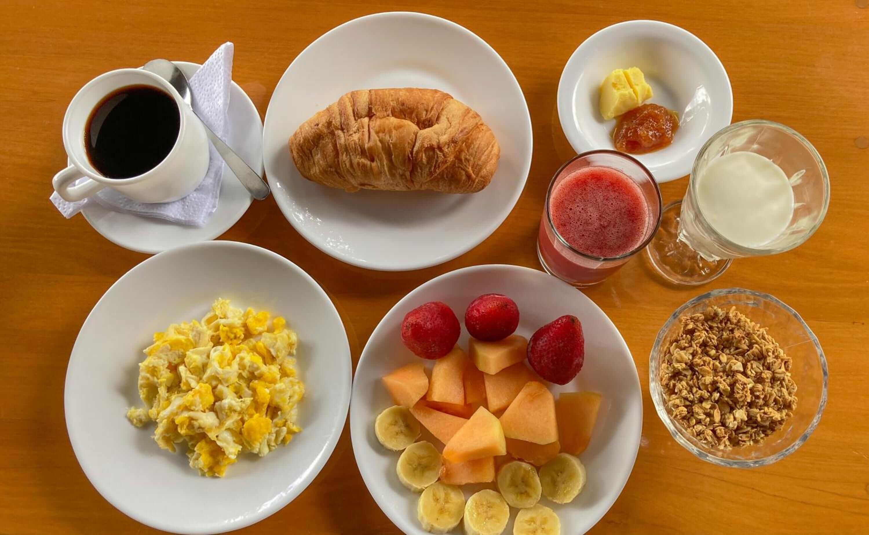 A breakfast table with scrambled eggs, a croissant, strawberries, banana slices, granola, a smoothie, and a cup of coffee.