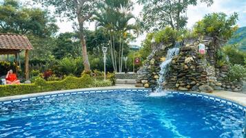 A swimming pool with a waterfall feature, surrounded by lush greenery and a gazebo.
