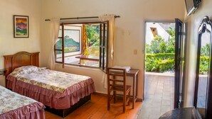 A room with two beds, a small table, and a window with a view of greenery. - Hostería Paraíso (Vilcabamba)