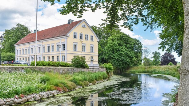 Ein großes, gelbes Gebäude mit rotem Dach, gelegen am Ufer eines ruhigen Flusses mit Spiegelungen.