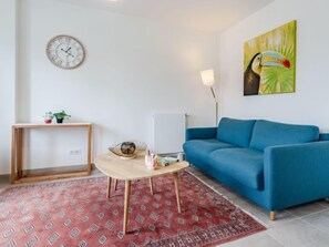 A living room with a blue sofa, a wooden coffee table, a red patterned rug, a wall clock, a small table with plants, and a painting of a toucan.