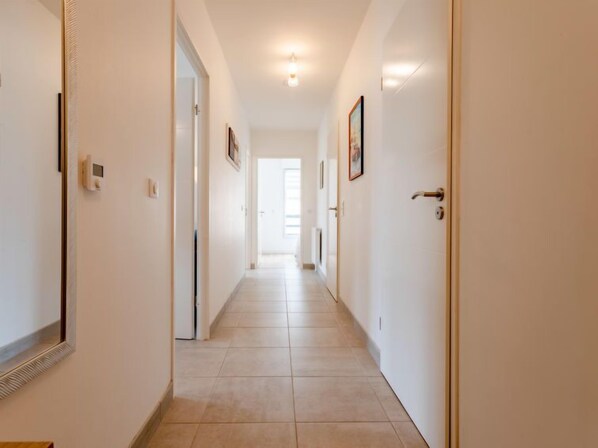 A long, narrow hallway with white walls and tiled flooring, leading to a bright room with a large window.